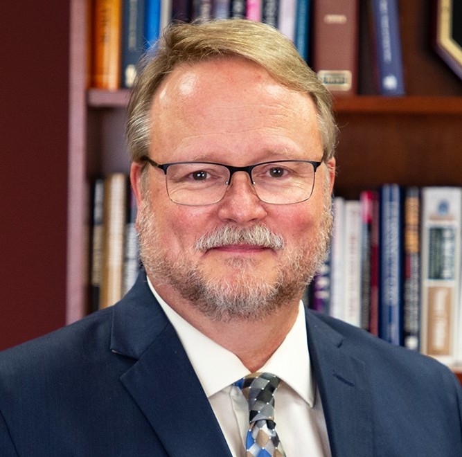 Portrait of a white man in a suit and glasses in front of bookshelves