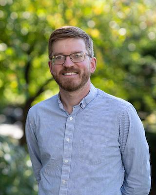 A man wearing a blue striped shirt in front of trees.