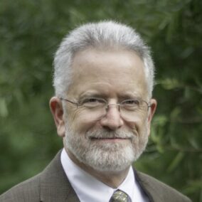 Man in light brown suit with glasses in a nature background.