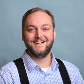 A white man in a blue check shirt and suspenders, smiling.