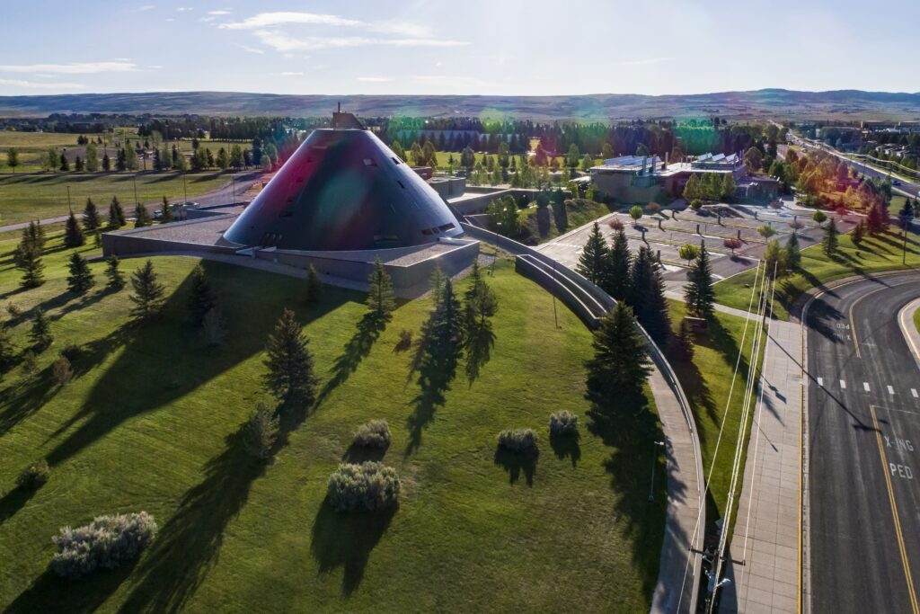 Aerial view of a large black, cone-shaped building surrounded by green lawns, evergreen trees, and nearby roads, with rolling hills stretching across the horizon under a clear blue sky.