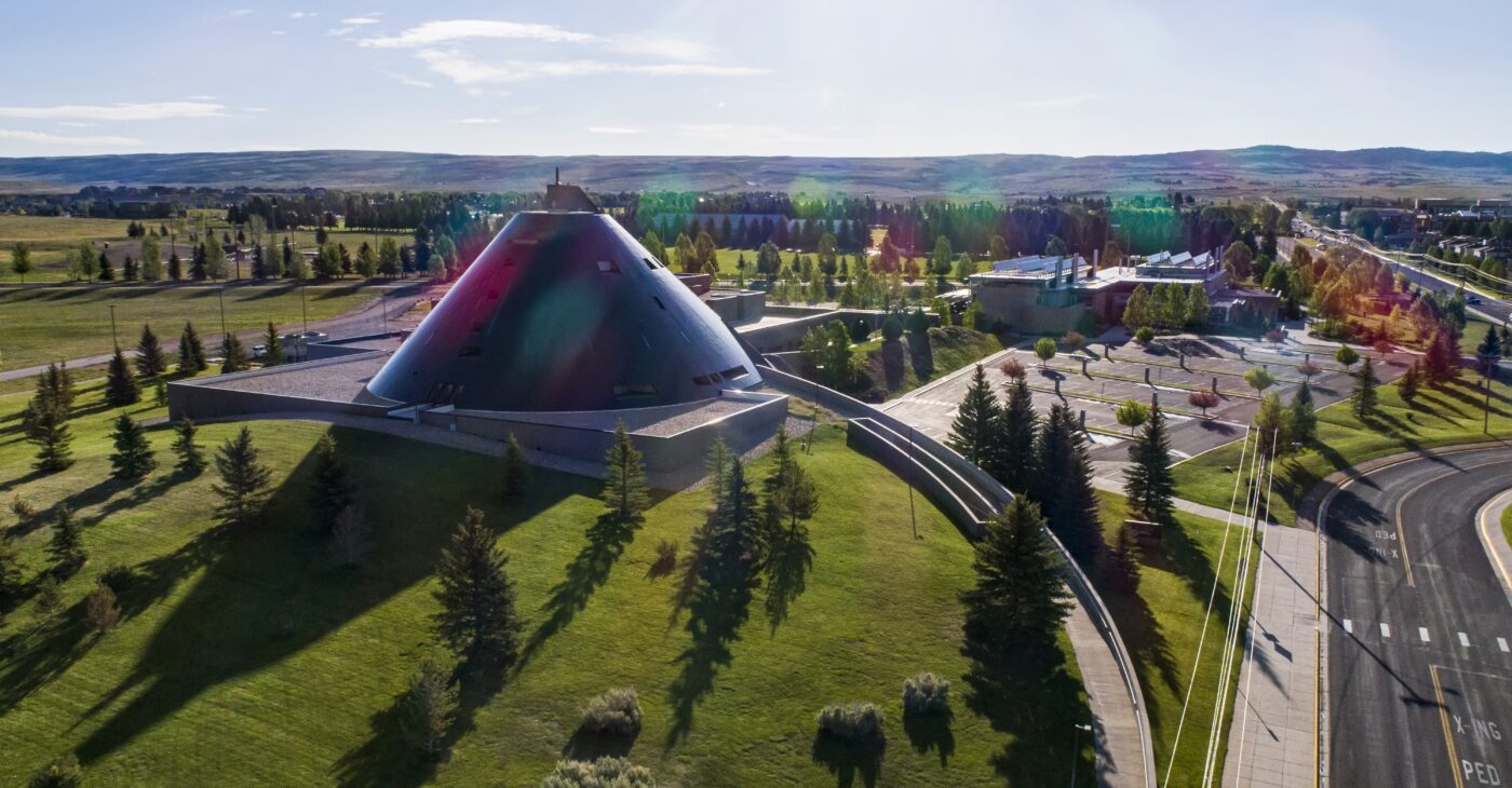 Aerial view of a large black, cone-shaped building surrounded by green lawns, evergreen trees, and nearby roads, with rolling hills stretching across the horizon under a clear blue sky.